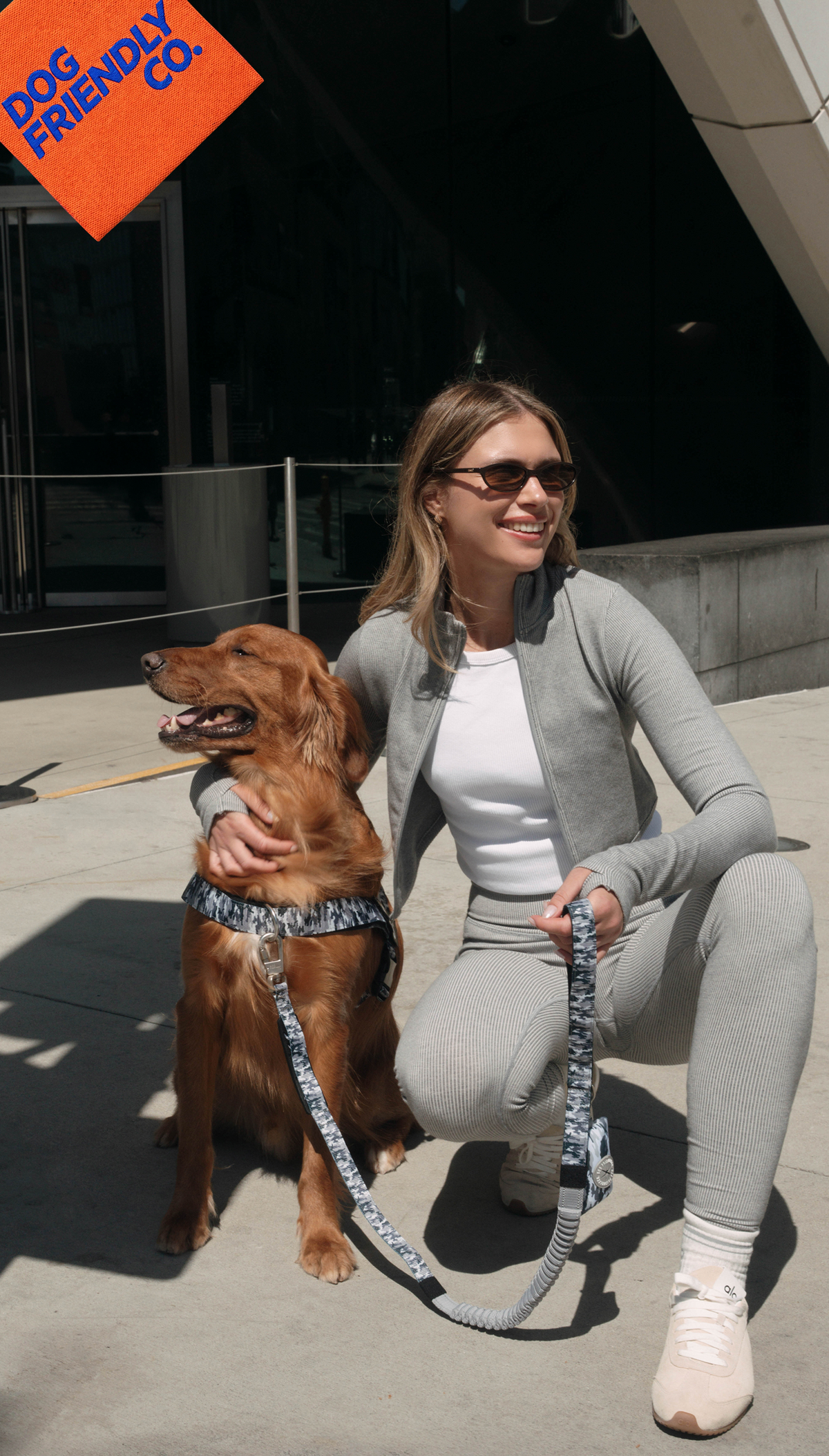 Woman kneeling beside a golden retriever on a sunny sidewalk